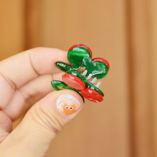Hand holding a cherry shaped hair clip with red and green berries against a wooden background. The claw is held open to reveal the transparent teeth inside.