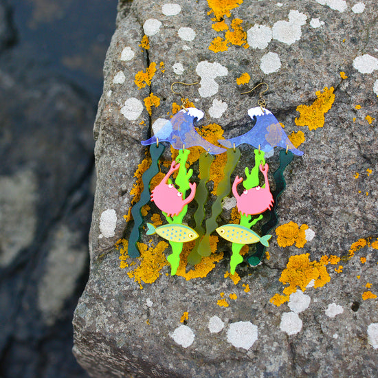 Very large earrings featuring a blue wave shape with green seaweed shapes dangling from it. There is also a yellow fish and a coral pink crab hanging from the seaweed. The background is a rock near the sea covered in moss.