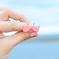 A close-up of a hand holding a pair of pink crab-shaped stud earrings against a soft, blurred blue background.