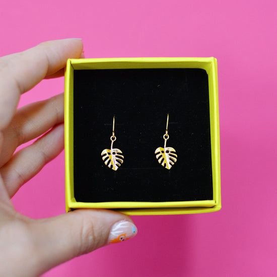Gold plated monstera leaf earrings in a yellow box with black velvet padding, held by a hand against a pink background