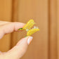 Hand holding a small yellow and green pineapple hair clip against a wooden background. The claw is held open so you can see the transparent teeth inside.