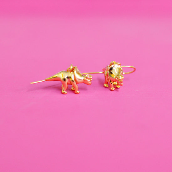 Gold plated triceratops shaped earrings on a pink background