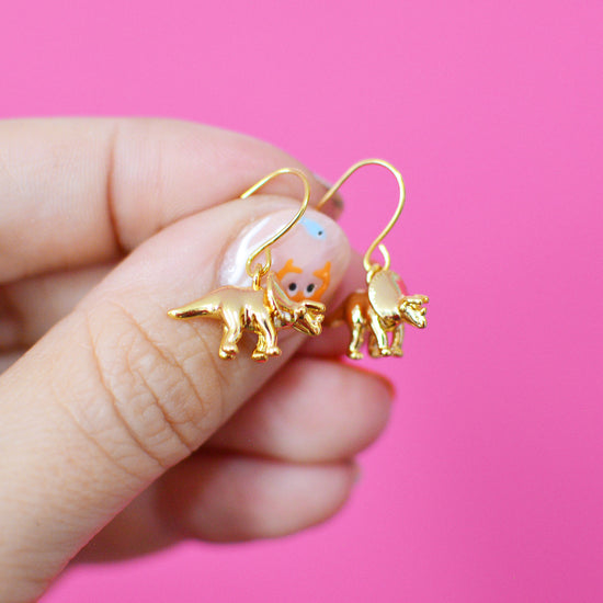 Gold plated triceratops dinosaur shaped dangle hook earrings held between fingers against a pink background