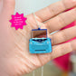 A hand holding a miniature blue typewriter pendant necklace with a tiny mirror where the paper would be inserted, suspended from a silver chain. A pink starburst graphic in the top left reads 'Do your lipstick in the tiny mirror!'.
